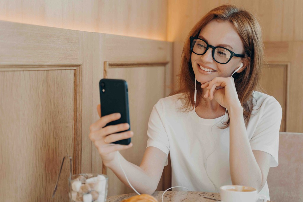 Smiling happy woman wearing eaphones and using her smartphone in cafe