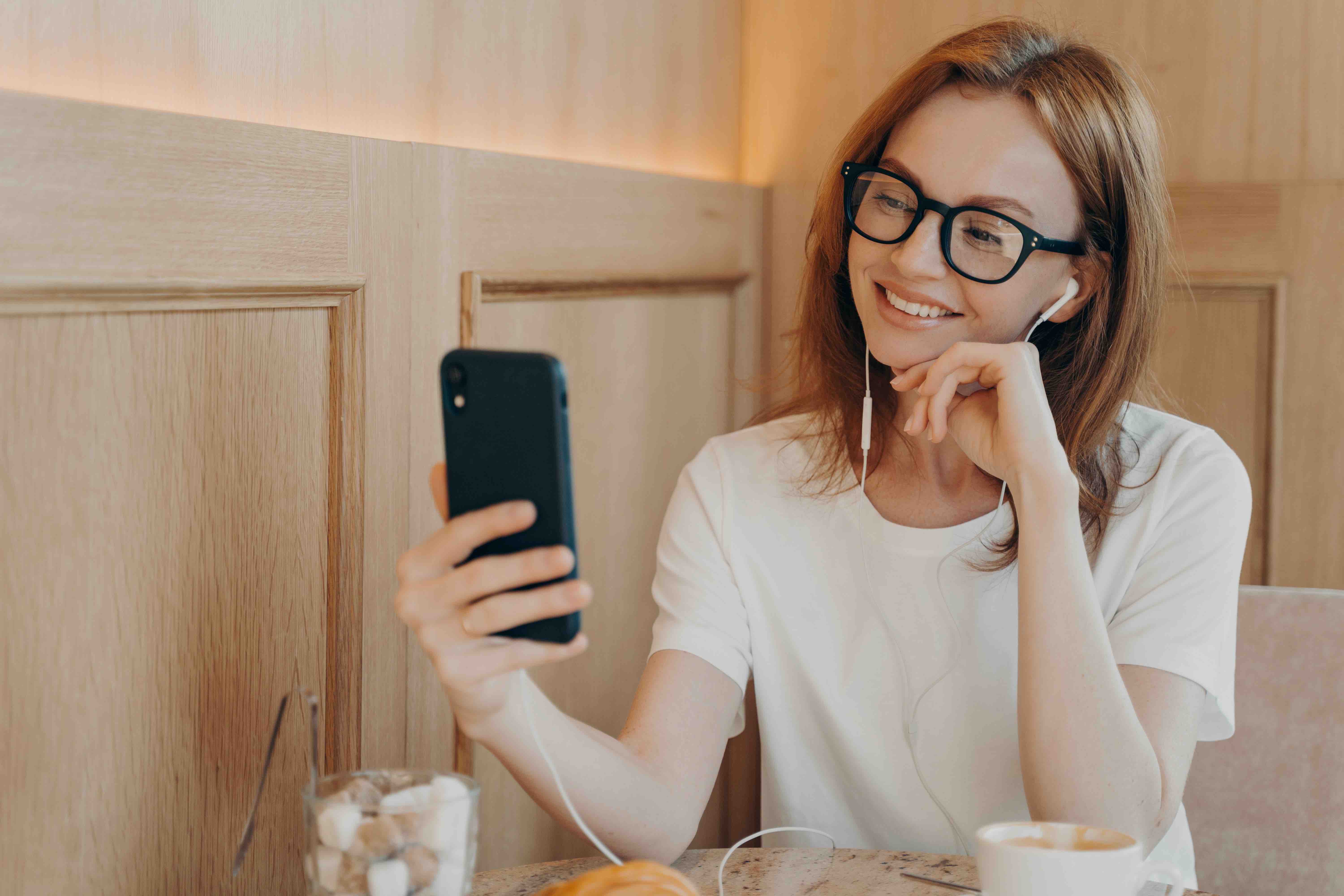 Smiling happy woman wearing eaphones and using her smartphone in cafe