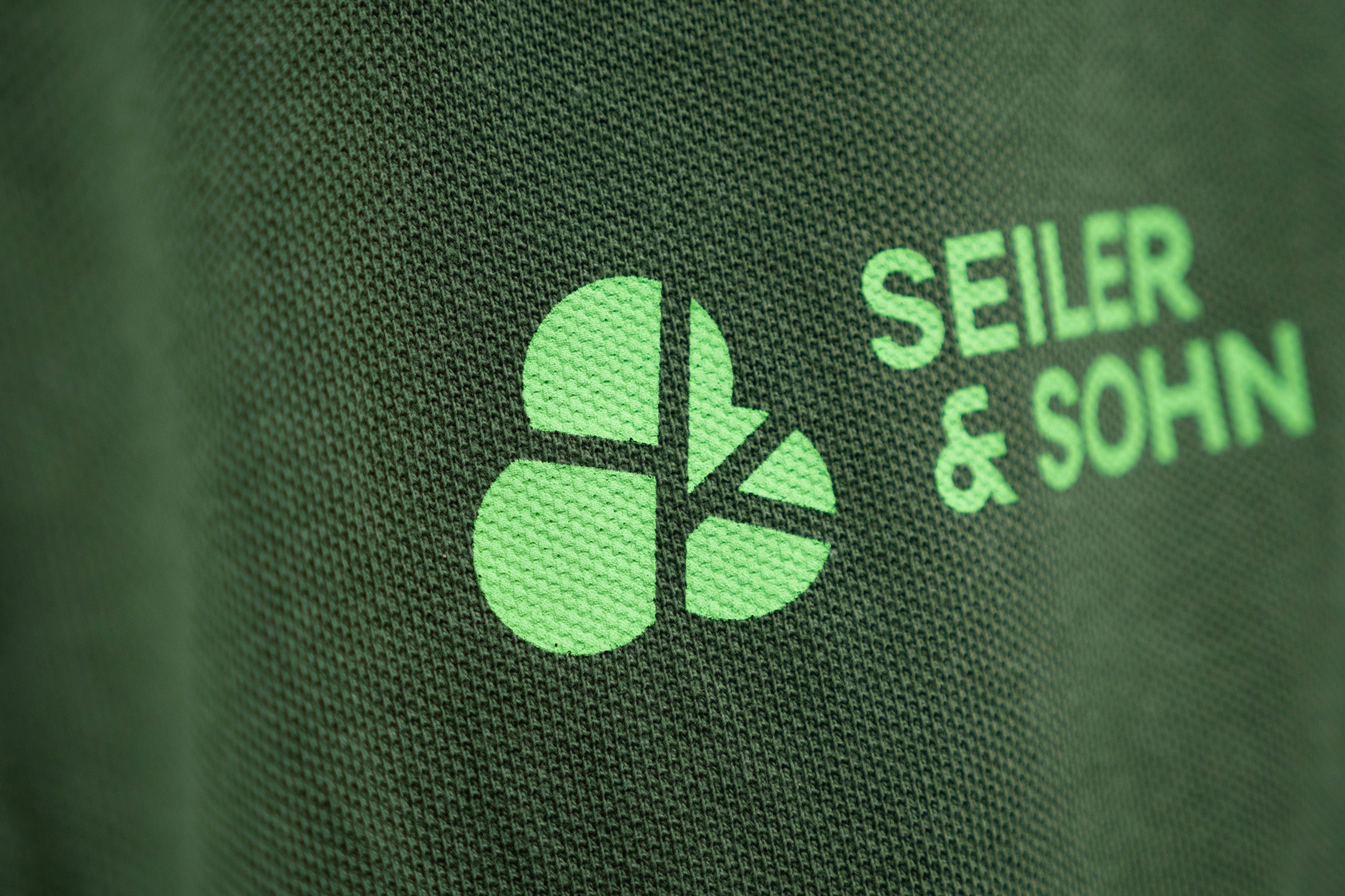 Nahaufnahme eines dunkelgrünen Textilstoffs mit dem Seiler & Sohn Logo in leuchtendem Lime-Grün – stilisiertes Kleeblatt-Icon und Schriftzug SEILER & SOHN.