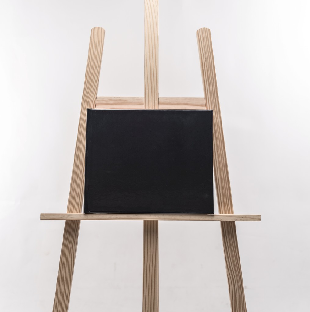 Fine Art wedding album with black leather cover and red gilded edges — photographed on a minimalist wooden easel. Album de mariage Fine Art à couverture noire et tranches rouges dorées — photographié sur un chevalet en bois minimaliste.