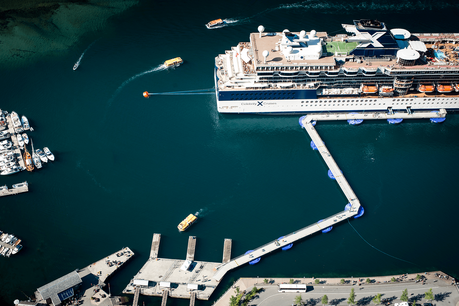 Aerial view of a cruise ship docked with the SeaWalk floating pier system extending from shore, with small boats navigating the harbor around the vessel.