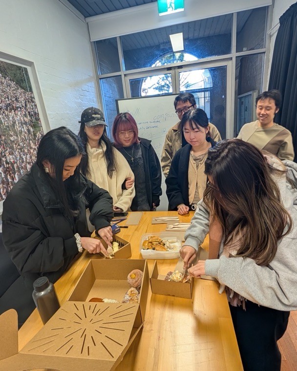 Students gathered around a long wooden table sharing takeaway food boxes in a UNSW Art & Design studio with a blue timber ceiling and stained-glass window.