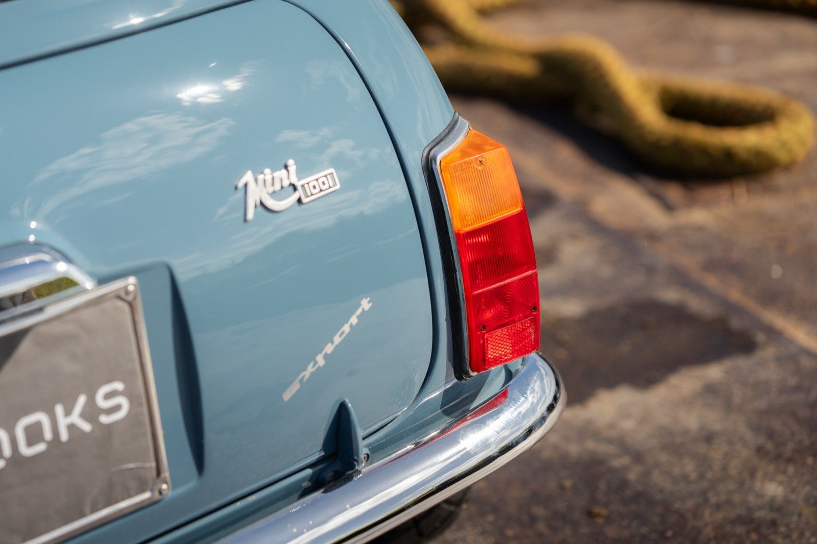 Close-up of a vintage blue car's rear, showcasing the tail light and part of the bumper on a sunny day.