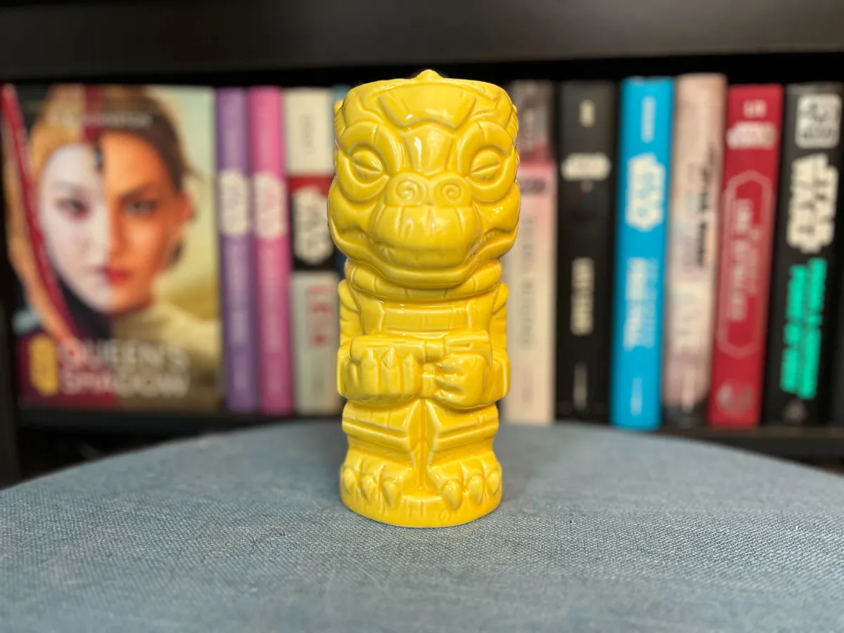 A Geeki Tiki mug in the shape of Bossk the bounty hunter on a blue surface in front of a blurred Star Wars bookshelf