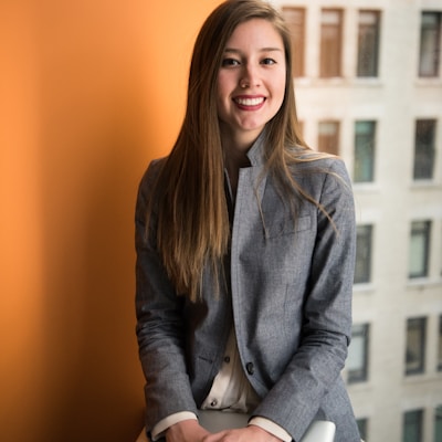 A person with long hair sits on a white chair, smiling, in front of an orange wall and a window with a city building backdrop.