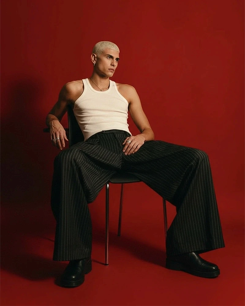 A stylish individual in a white tank top and pinstripe trousers sits against a bold red background, exuding a modern and confident vibe.