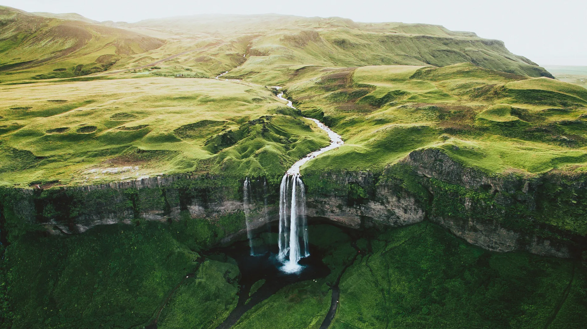Aerial of a waterfall dropping from a green clifftop onto a dark plain