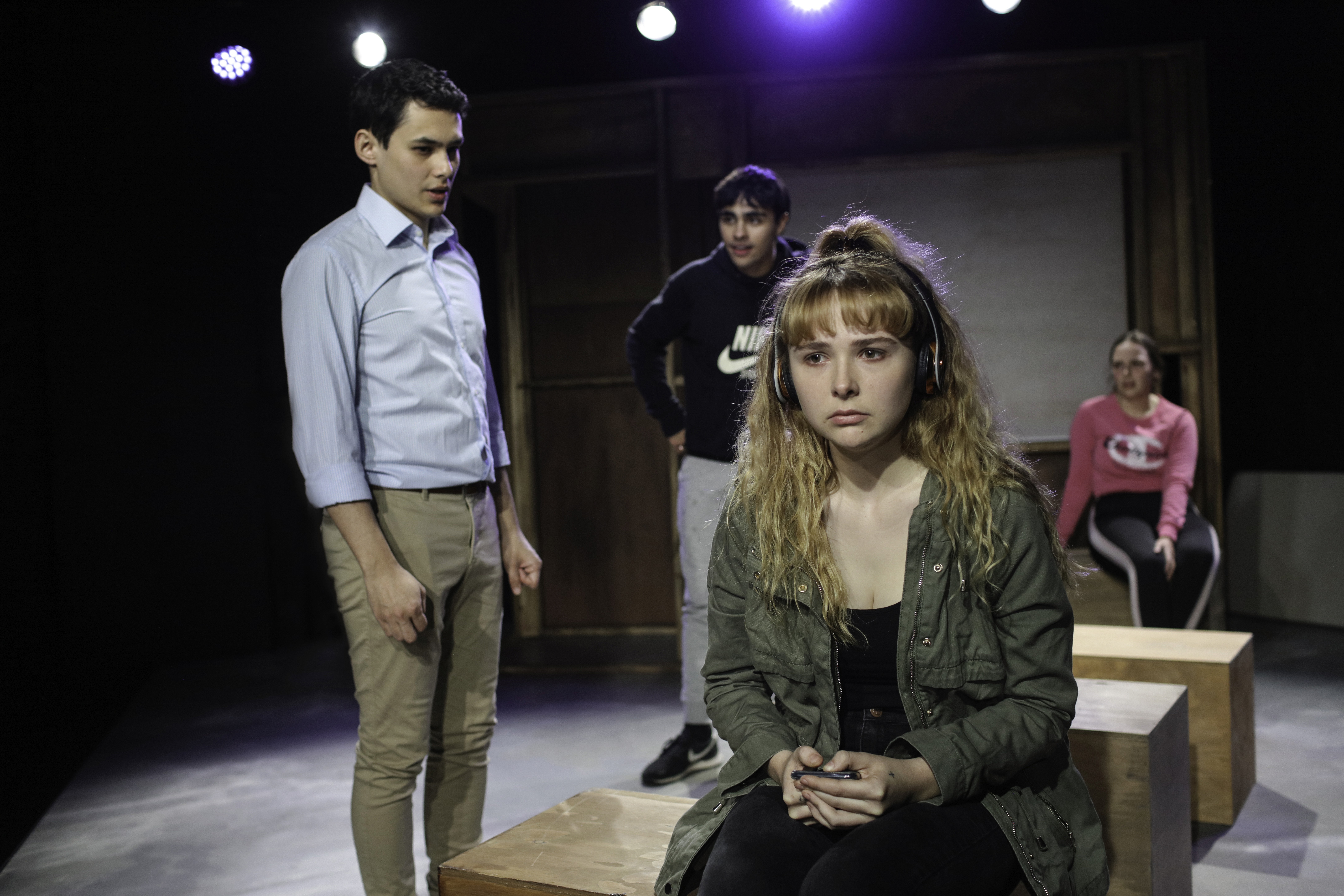 A teenage girl wearing headphones sits on a block, looking sad and withdrawn. Three other performers stand behind her, watching with concern in a stark, tense scene.