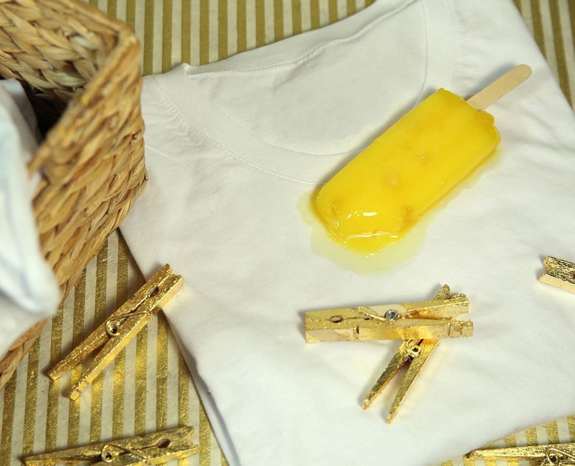 A yellow popsicle rests on a white t-shirt, its stick protruding from the top. A pool of yellow liquid is visible on the shirt near the popsicle. Four gold clothespins are arranged on the white fabric beneath the shirt. A woven basket is partially visible in the upper left corner. The background consists of yellow and white striped fabric.