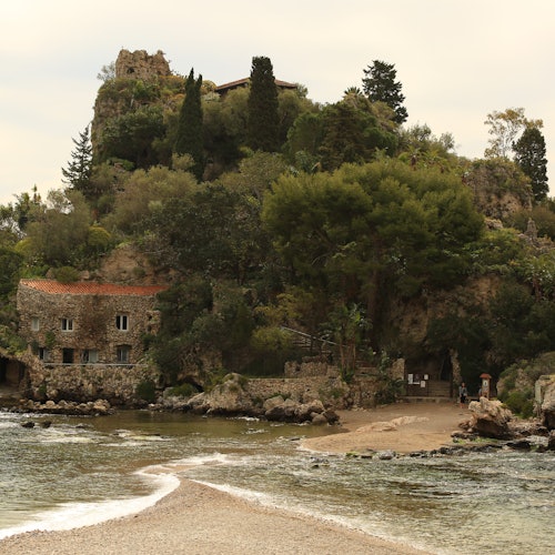 Une maison en pierre avec un toit rouge entourée d'arbres près d'un rivage rocheux et d'une étendue d'eau, avec une colline et des ruines en arrière-plan.