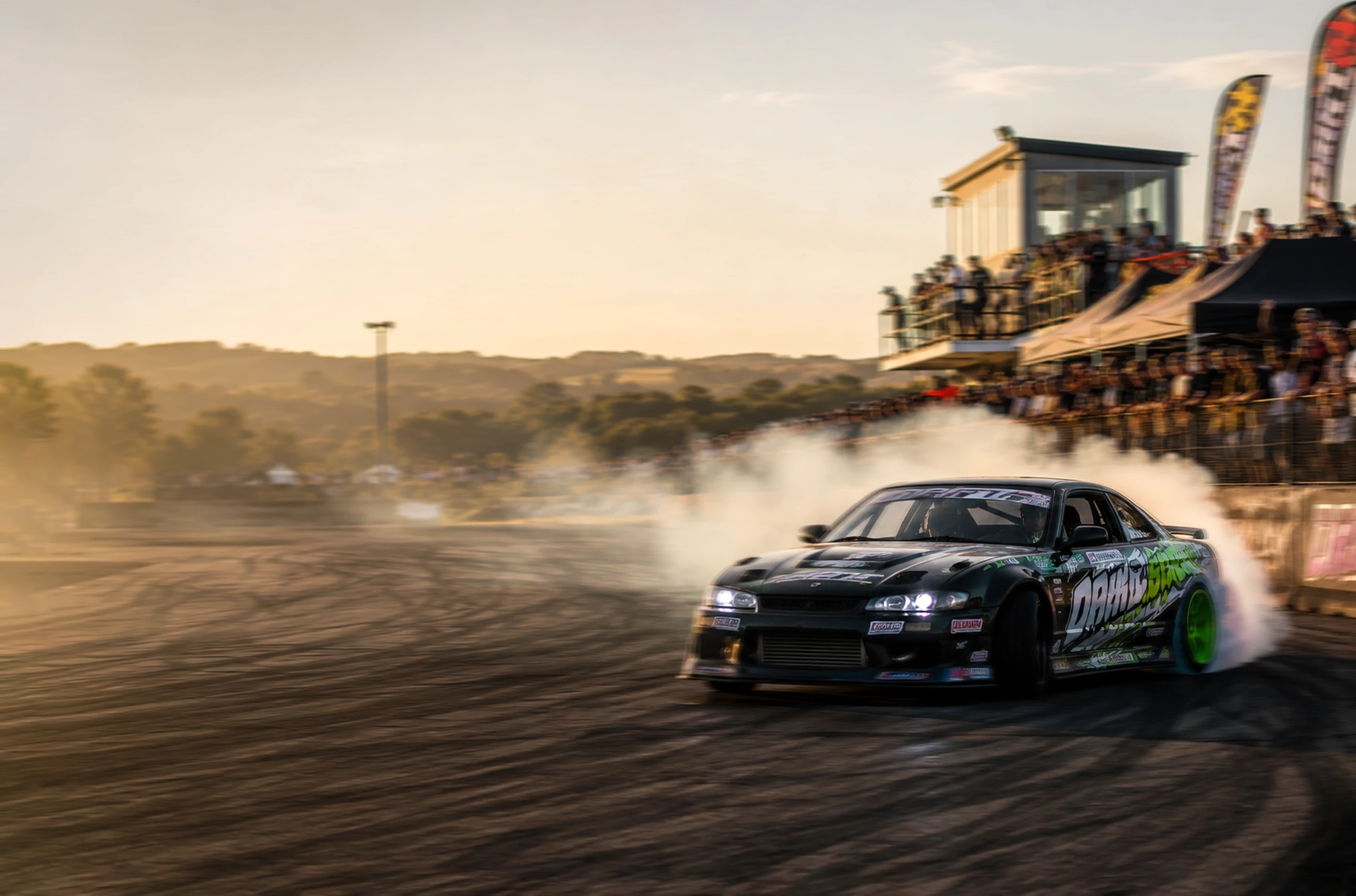 a drift car slides with smoking tires in front of a packed crowd at sunset