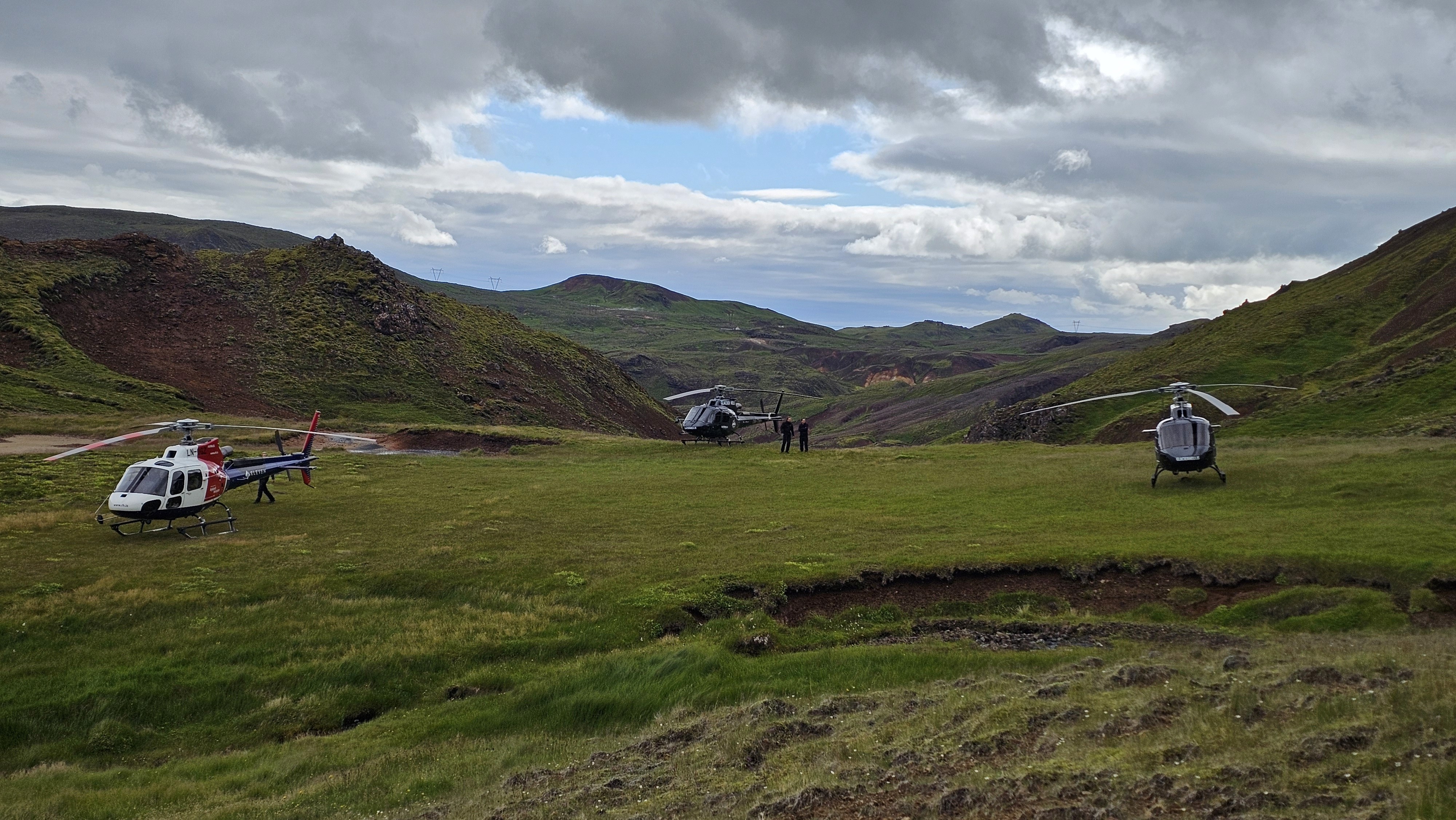 Three helicopters are parked on a grassy field surrounded by green and red-tinged hills under a partly cloudy sky. A few people stand near the center helicopter, indicating possible exploration or a remote landing in Iceland’s rugged highlands.
