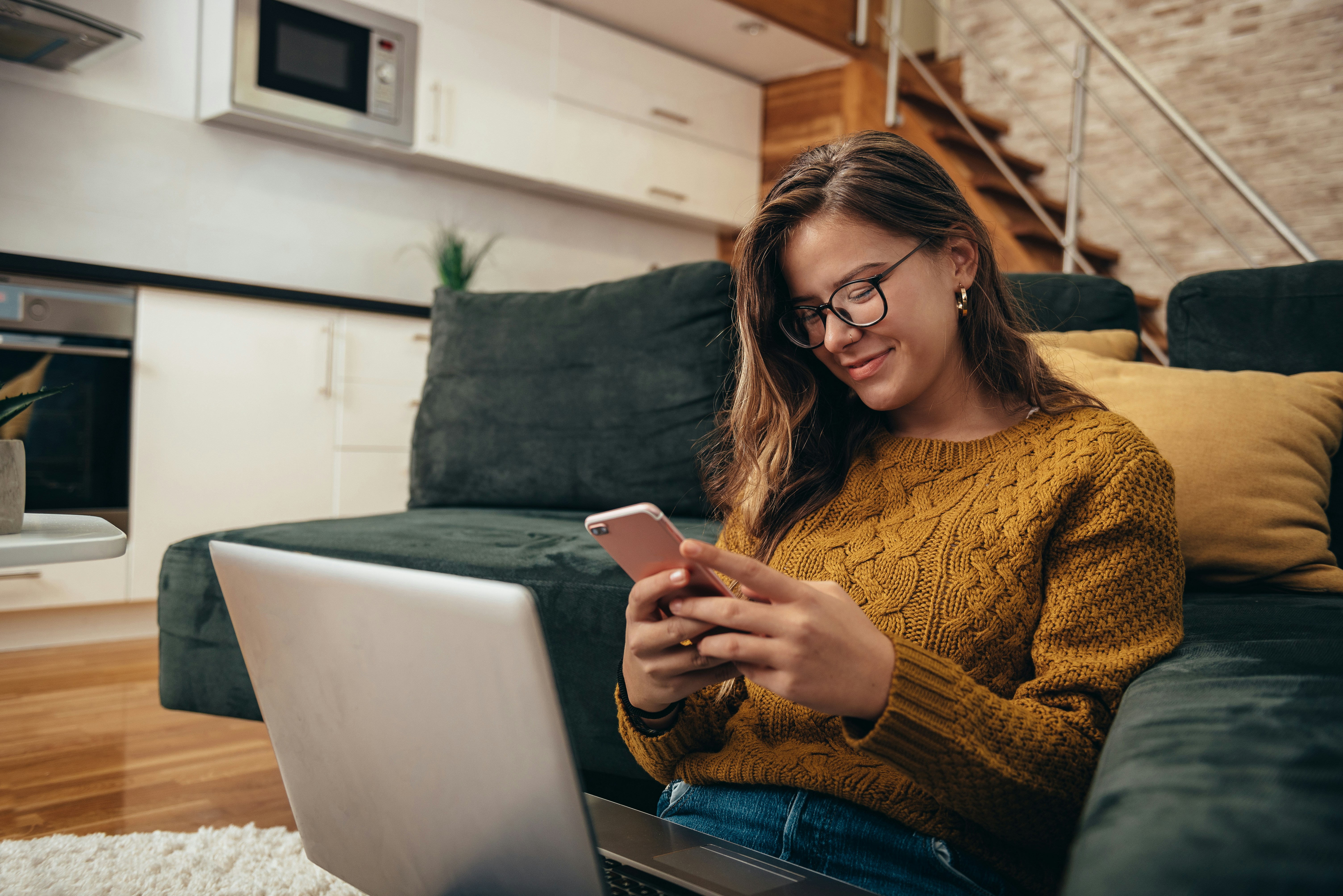 A woman in a brown sweater sits on a couch, looking at her phone while using a laptop on a table.