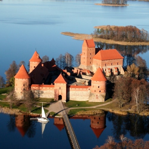 Aerial view of a castle with red-roofed towers surrounded by a lake, connected to land by a wooden bridge and a sailboat nearby.