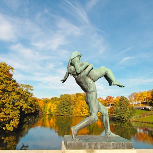 Бронзовая статуя фигуры, несущей другую, над озером с осенними деревьями и синим небом на заднем плане.