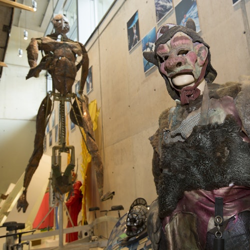 Mostra con grandi sculture umanoidi e manichini che indossano maschere e costumi artistici dettagliati in uno spazio industriale.