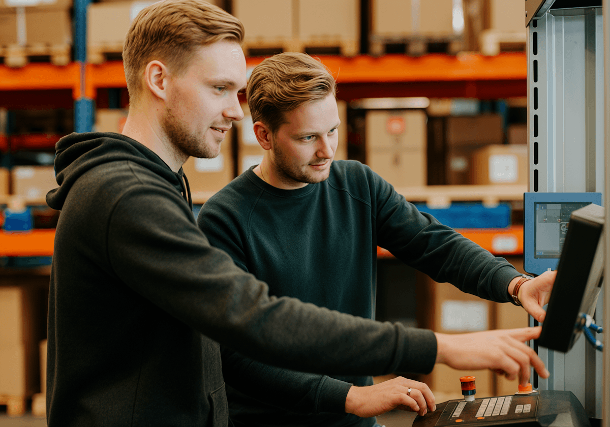 Two individuals collaborating on a task in a warehouse, focusing on a control panel with shelves and equipment in the background.