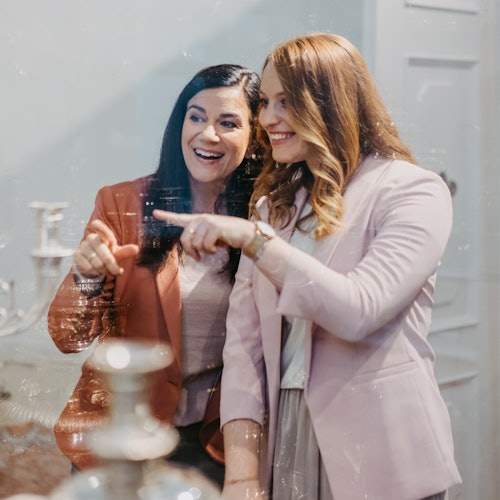 Zwei Frauen in Blazern lächeln und zeigen auf etwas vor ihnen, mit verschwommenen Objekten im Vordergrund.