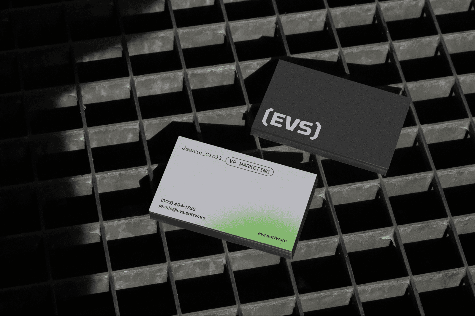 Two EVS business cards on metal grating: a black card with the “(EVS)” logo and a grey card for Jeanie Croll, VP Marketing, with a green gradient and evs.software