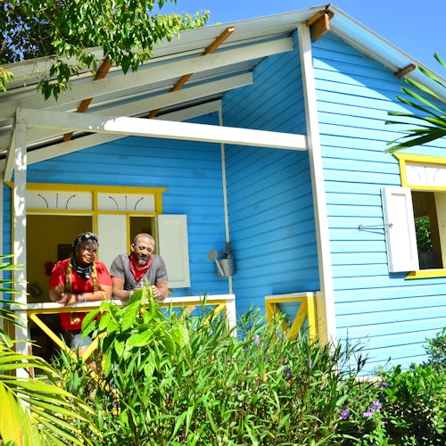 Dos personas en el porche de una casa vibrante azul y amarilla, rodeada de exuberante vegetación bajo un cielo azul claro.