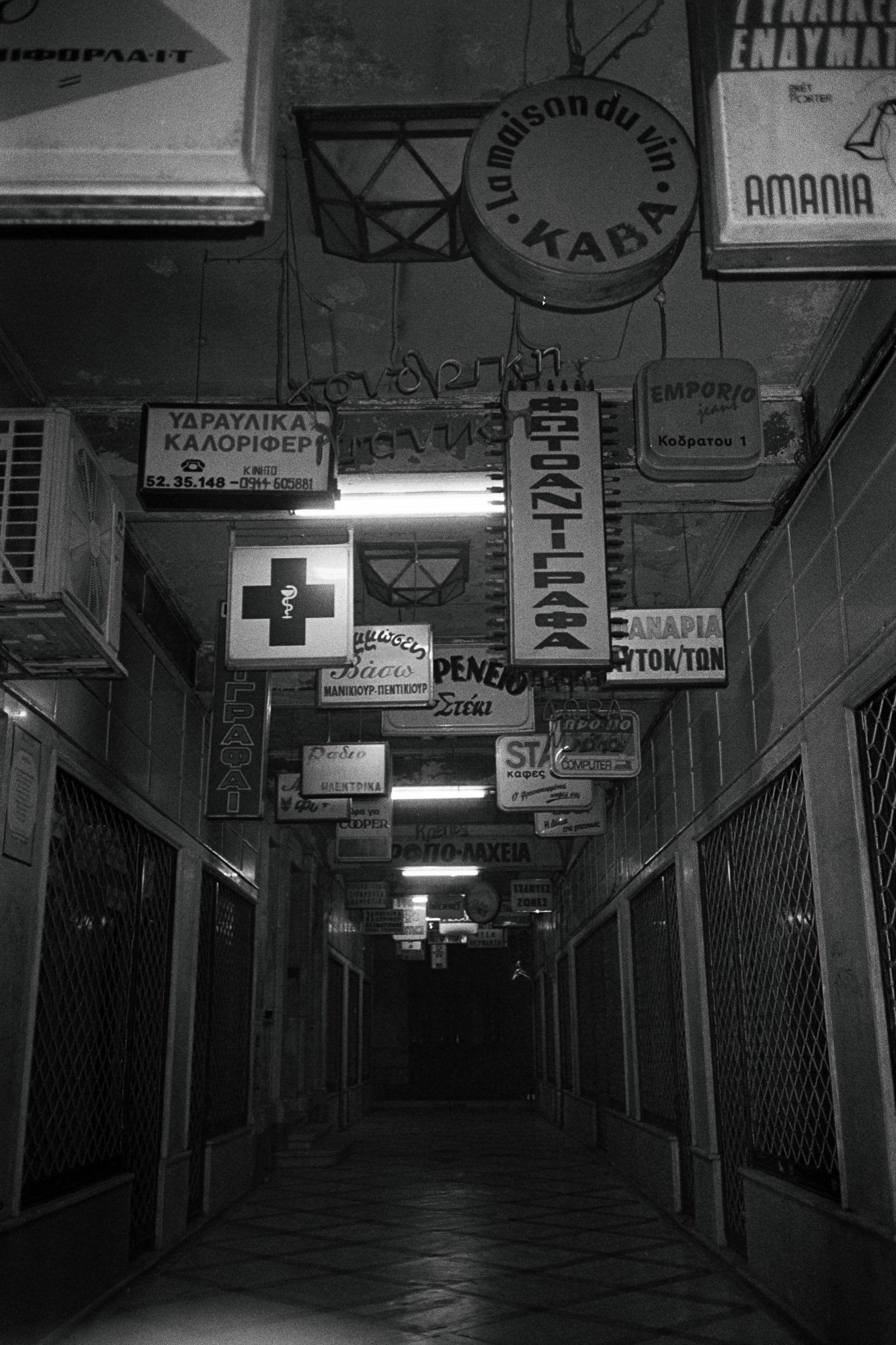 A dimly lit, narrow corridor lined with vintage signs in various languages hangs overhead, creating an atmospheric scene of an old shopping arcade or passageway, with visible text and decorative elements.