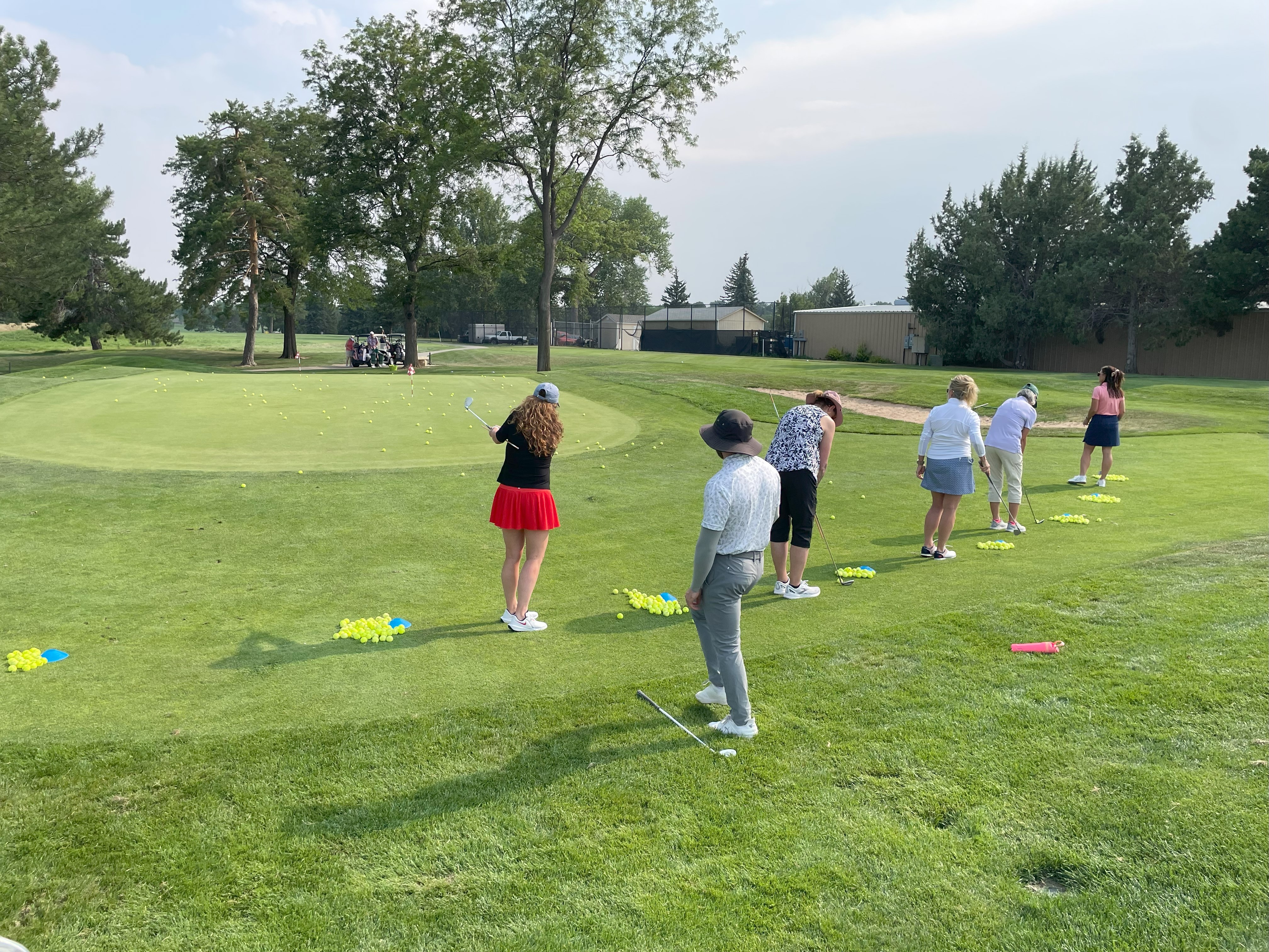 A group of students at the edge of a green, chipping balls towards net targets.