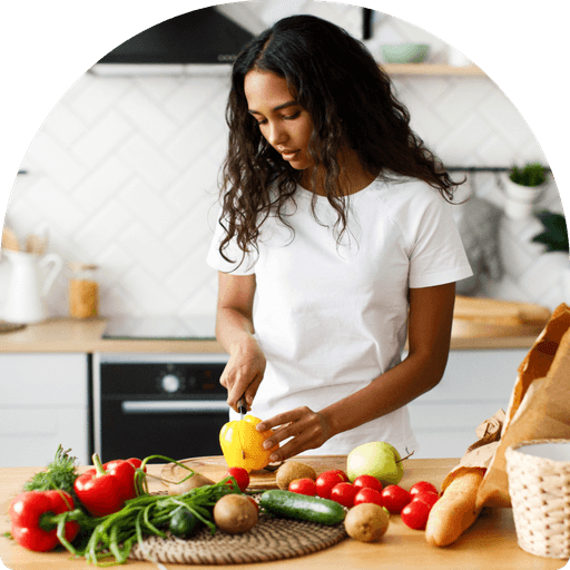 In a bright kitchen, a woman in a white t-shirt slices a yellow bell pepper as part of her nutrition plan. The wooden countertop, symbolizing wellness, is adorned with fresh vegetables like tomatoes, cucumbers, and mushrooms, alongside a loaf of bread.