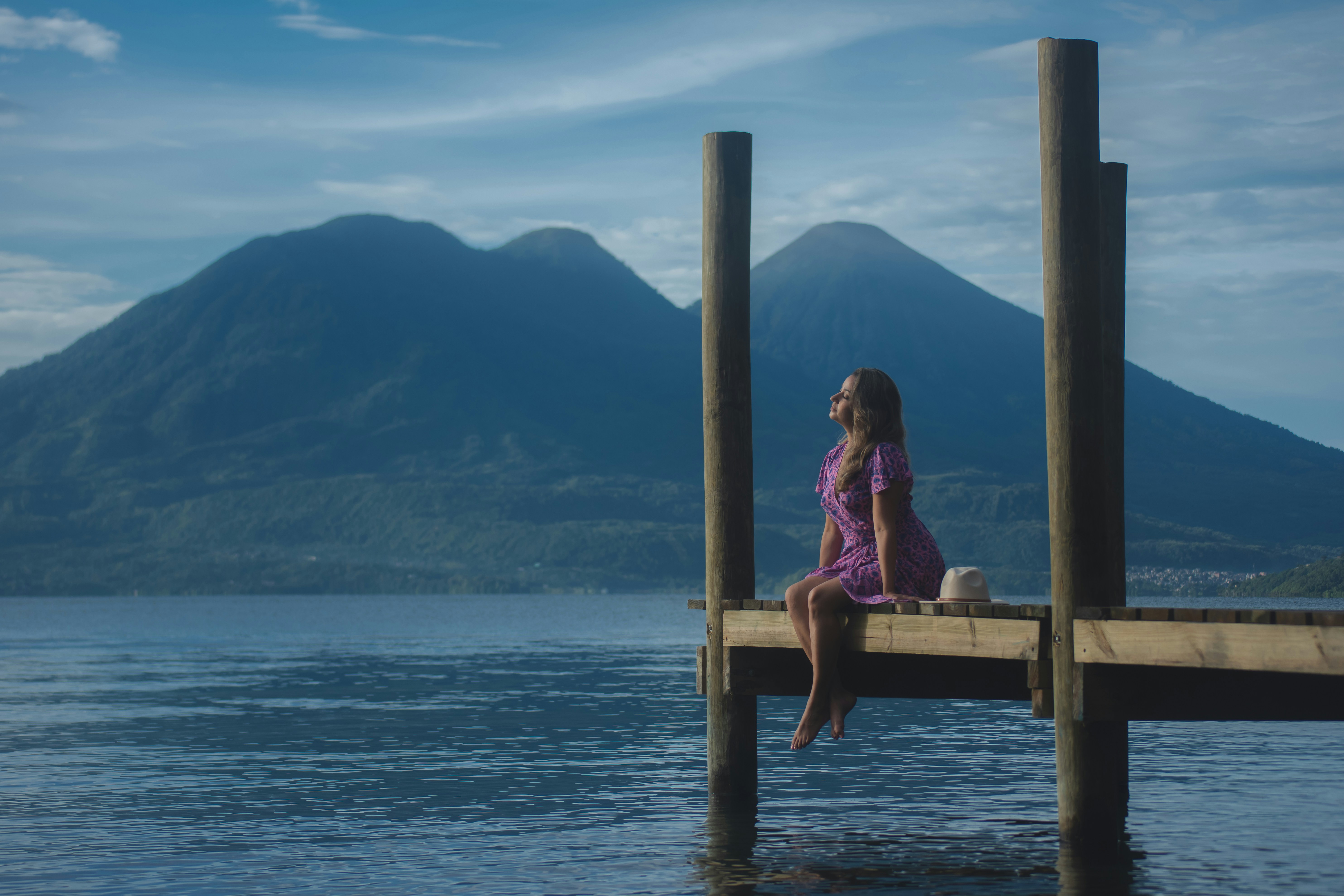 A woman in a purple dress sits peacefully on a wooden dock overlooking a serene lake, with two majestic volcanoes under a partly cloudy sky in the background, creating a tranquil scenic view.
