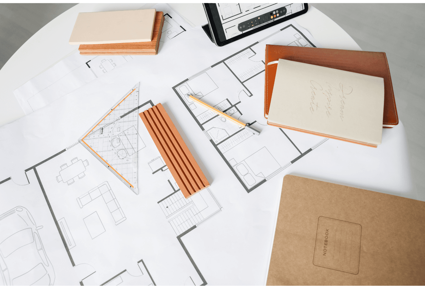 Architectural floor plans on a desk surrounded by drafting tools, notebooks, and wood material samples.