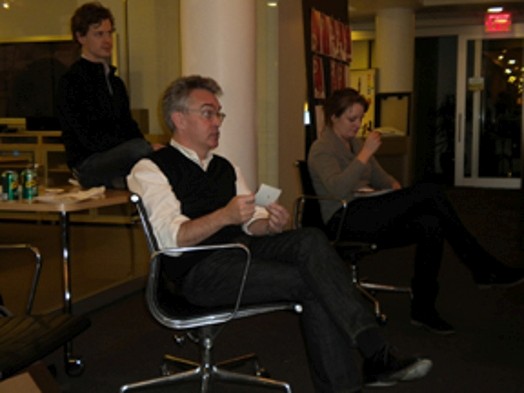 A photo showing a couple of people siting on chars and standing while listening to some lecture.
