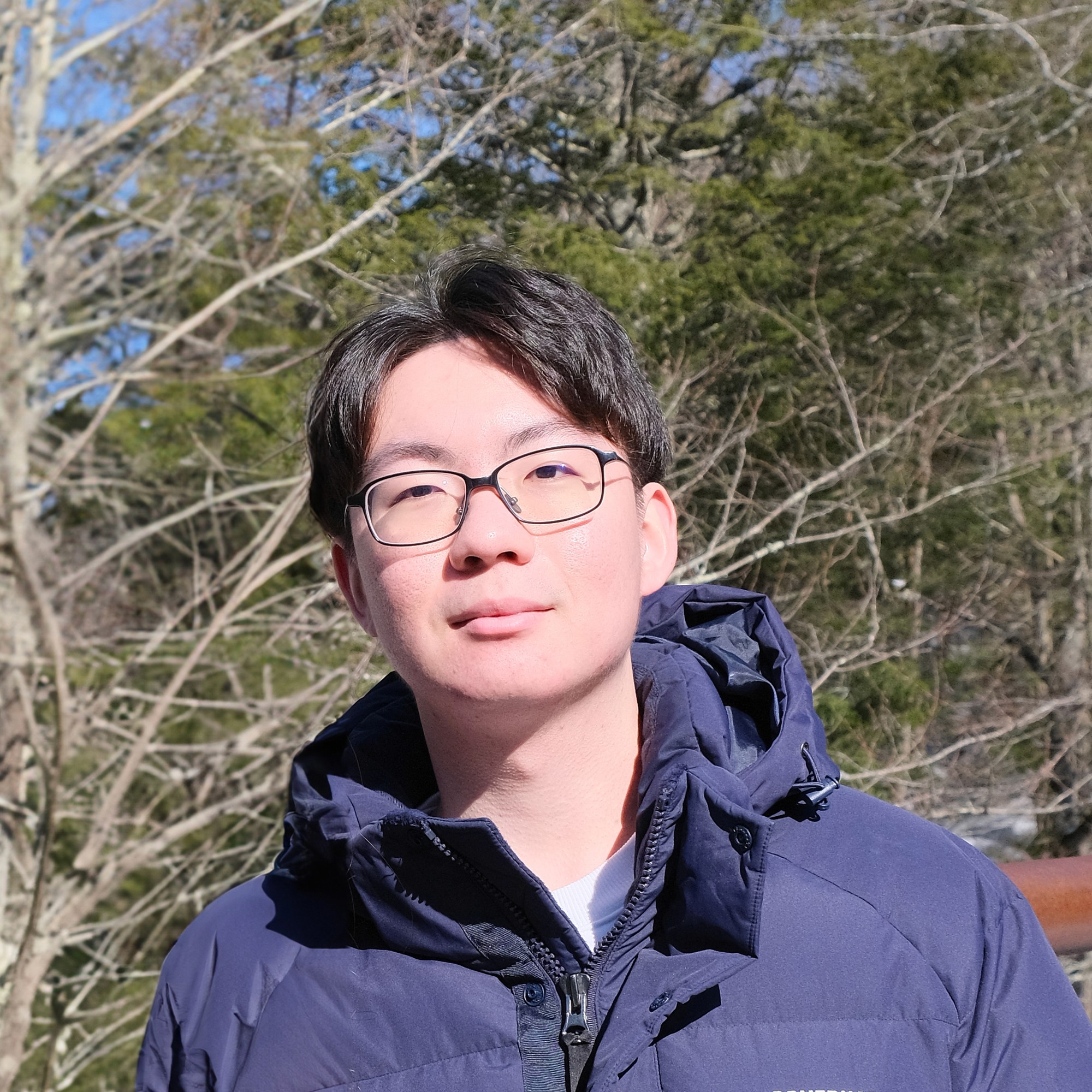 man looking at camera with glasses, black hair, and a blue hooded jacket