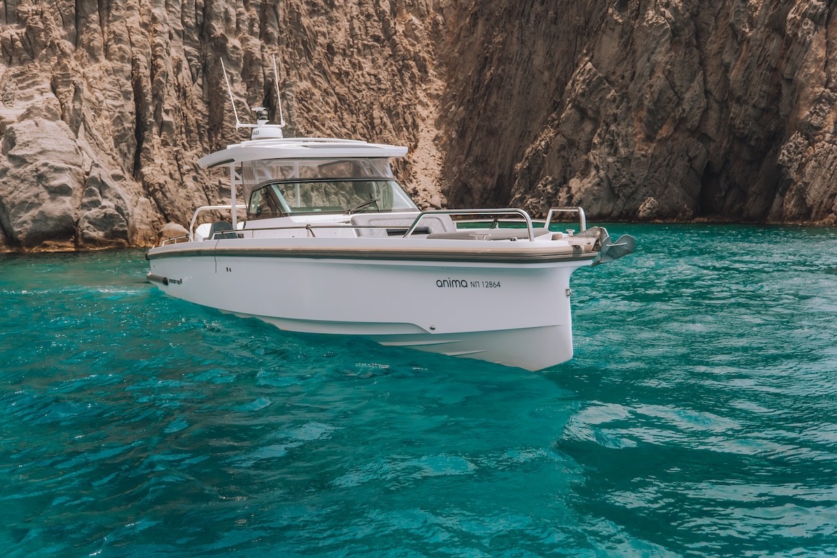 White Axopar 37 motor yacht anchored in crystal-clear turquoise waters beside dramatic layered rock cliffs in the Cyclades.