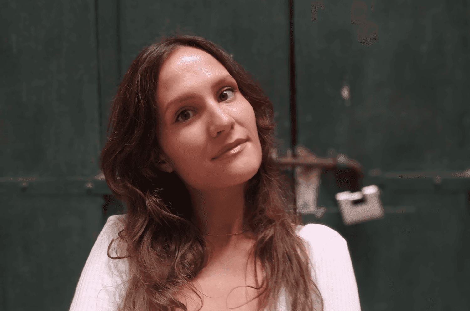 A person with long, wavy hair and a calm expression stands in front of a dark green metal door with a visible padlock.