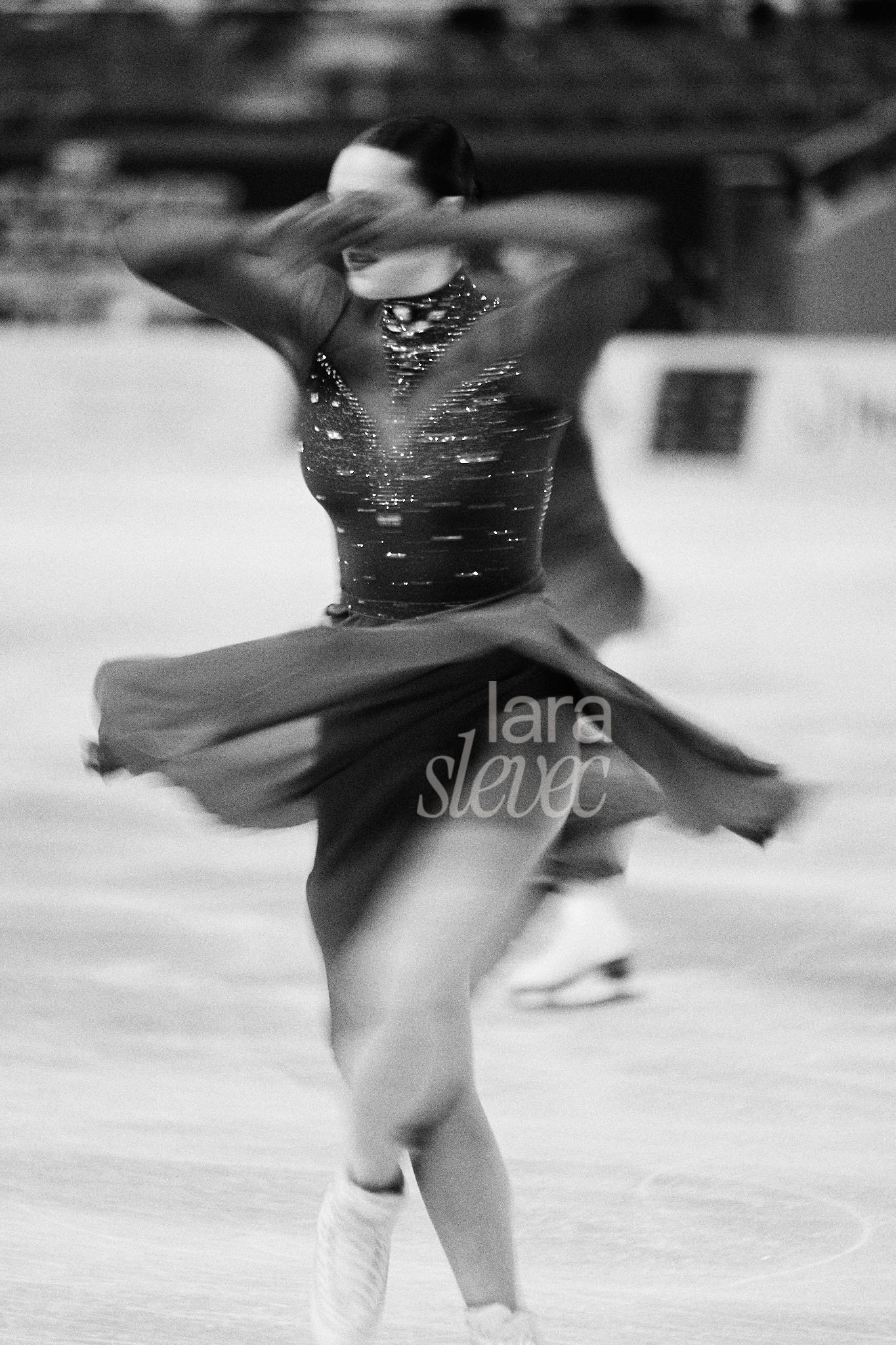 Schwarz-Weiß Bild im Analog-Stil. Eine Eiskunstläuferin in bewegungsunschärfe während sie eine Pirouette dreht. Die Hände hat sie vor ihren Augen. Team Ice Fire. ISU Synchronized Skating Championships 2026 in Salzburg. Fotografiert von Lara Slevec.