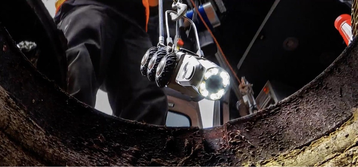 A large industrial facility with gray metal siding and a tall smokestack is in the background, while a white semi-truck towing a cylindrical tanker is parked in the foreground.