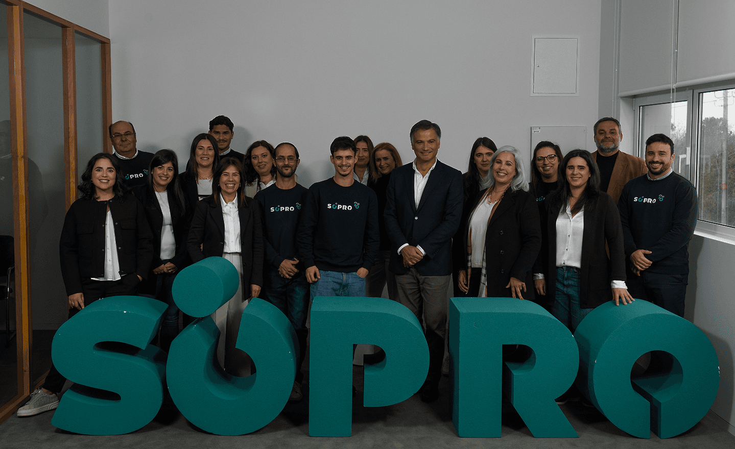 Grupo de pessoas sorrindo ao lado de grandes letras 3D formando a palavra  SRO, em um ambiente corporativo moderno, destacando a equipe da S/PRO na campanha de destaque na área de tecnologia e inovação.