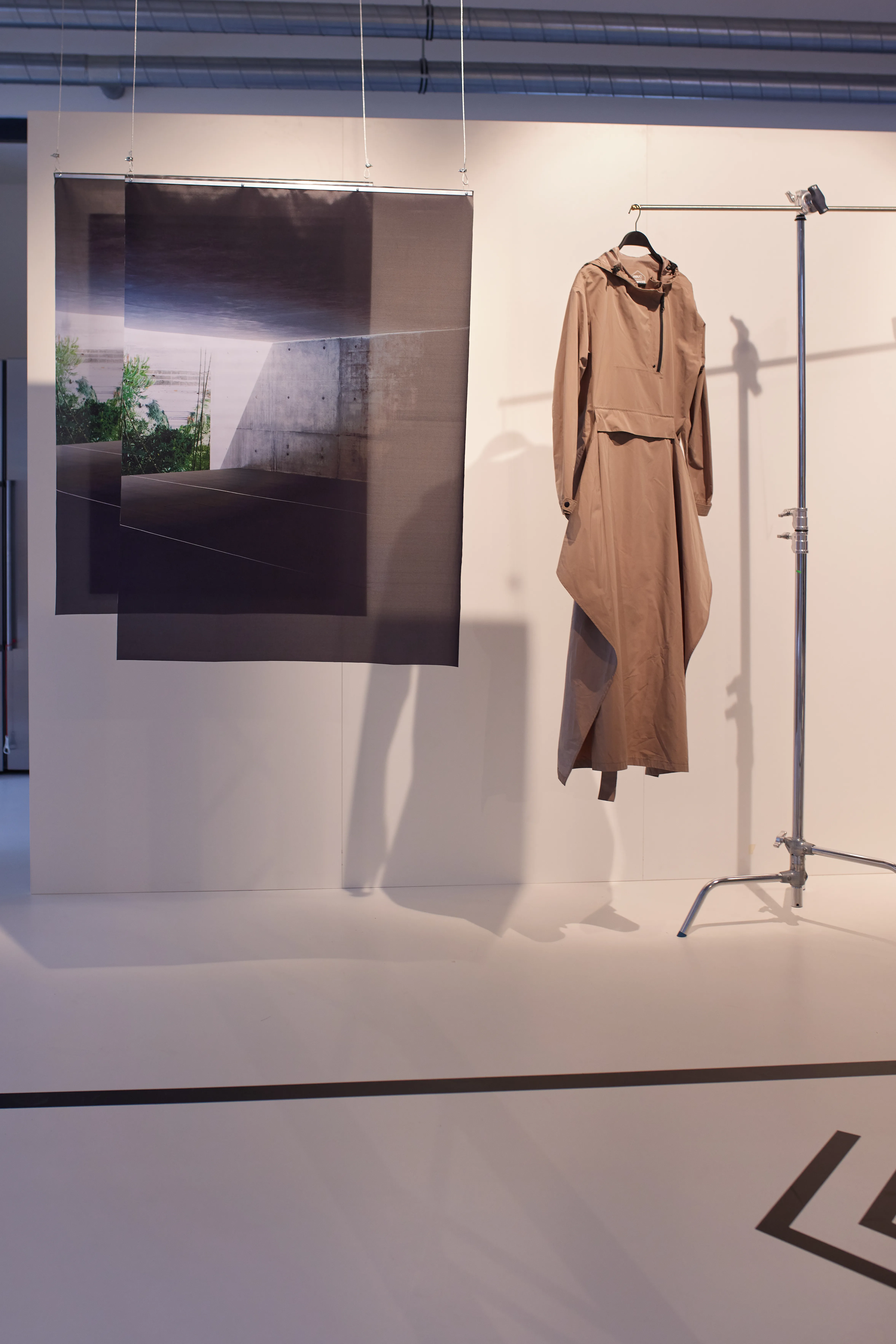 Camel-colored AGU rain poncho on a steel rail next to layered architectural photo prints in the white exhibition space.