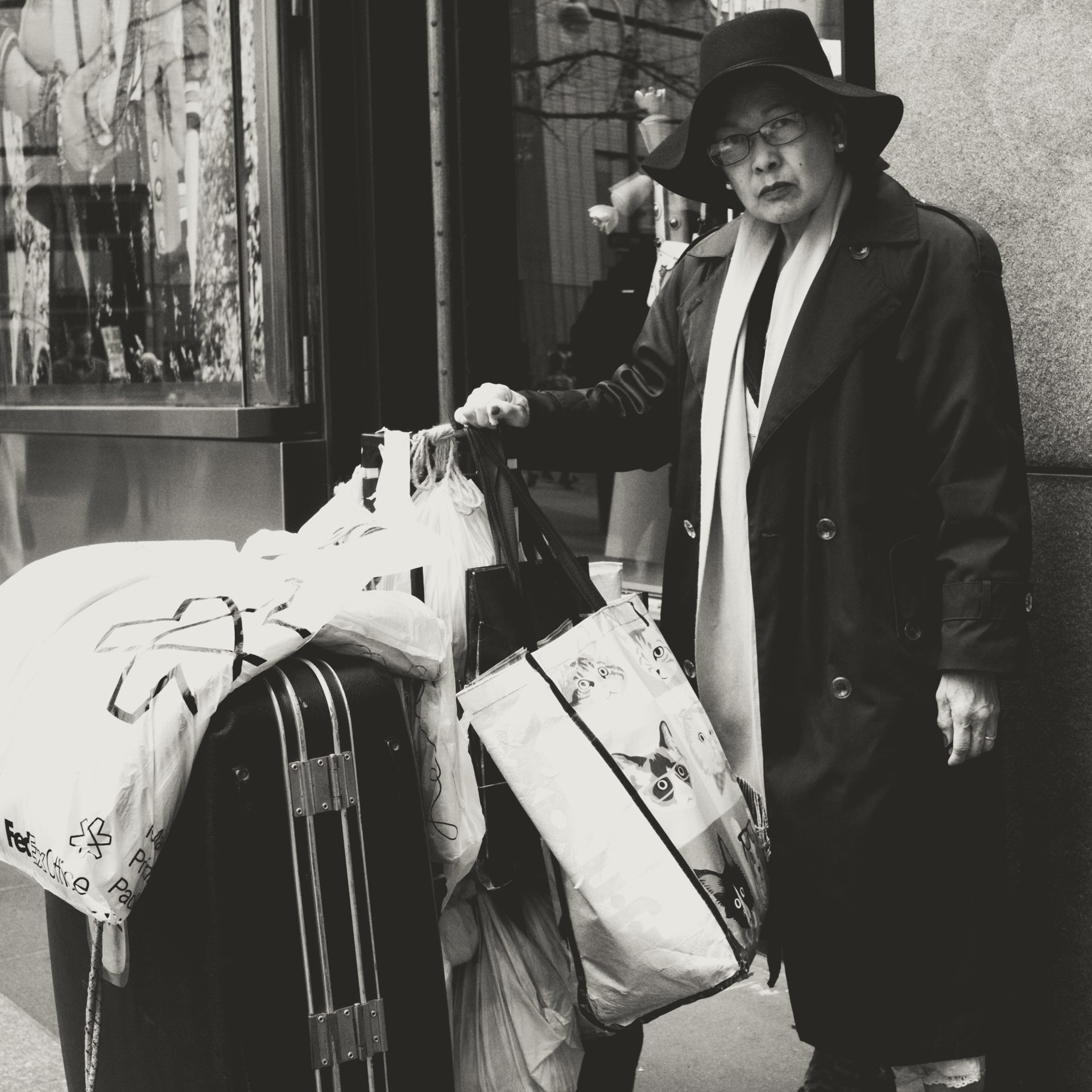 Black-and-white street photography of a traveler with luggage on a city sidewalk — candid urban portrait showing a person in a hat and scarf beside suitcases and shopping bags in front of a storefront; ideal for content about travel, urban life, street photography, and city portraits.