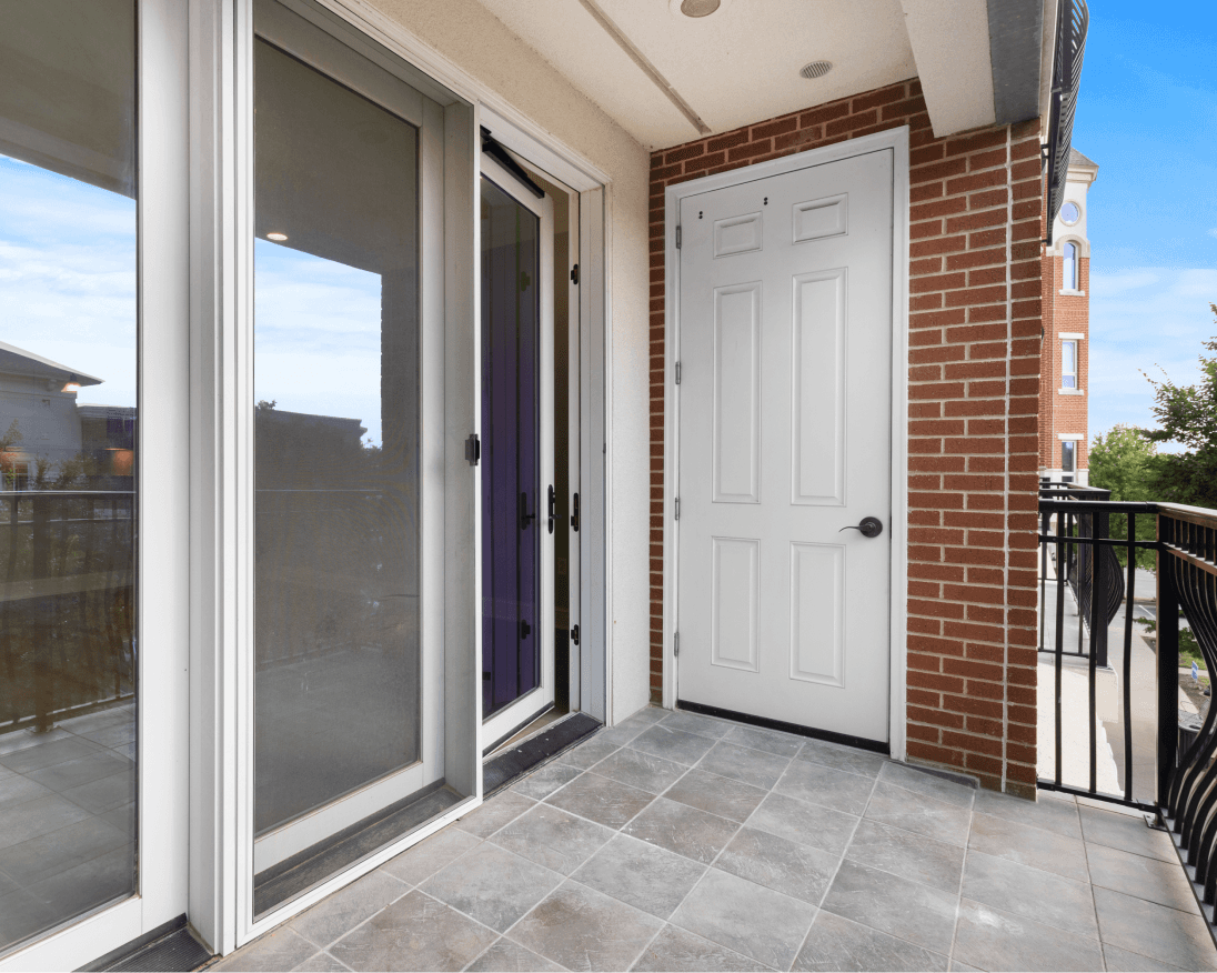 Tiled apartment balcony at The Gateway Village with sliding glass and white doors, brick walls, and a metal railing.