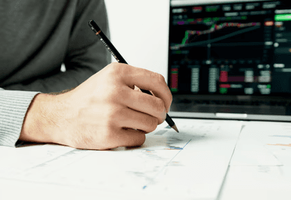 Person analyzing financial charts on paper with a laptop displaying stock data.