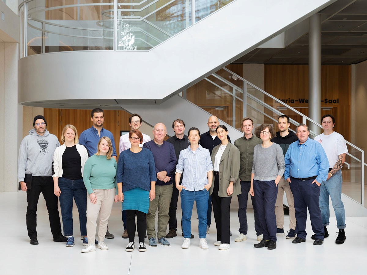Eine bunte Gruppe von Menschen steht in einem modernen Gebäude mit Treppen aus Metall und Glas und großen Fenstern zusammen.