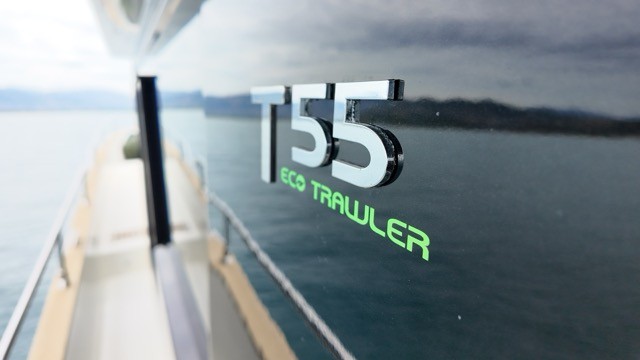 Close-up detail of the Cranchi T55 Eco Trawler model name on the hull side, with chrome three-dimensional T55 lettering and neon green Eco Trawler text against the dark superstructure glass, with the teak side deck and open sea visible beyond