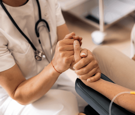 A healthcare professional in scrubs with a stethoscope is holding the hand of a patient, offering comfort and support. The patient’s arm is resting on a medical device, and an IV line is visible. The setting appears to be a medical facility.