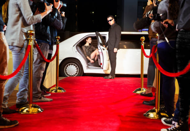 A man opens the door of a while limousing for a woman woman to steps out onto a red carpet, which is lined with photographers. 