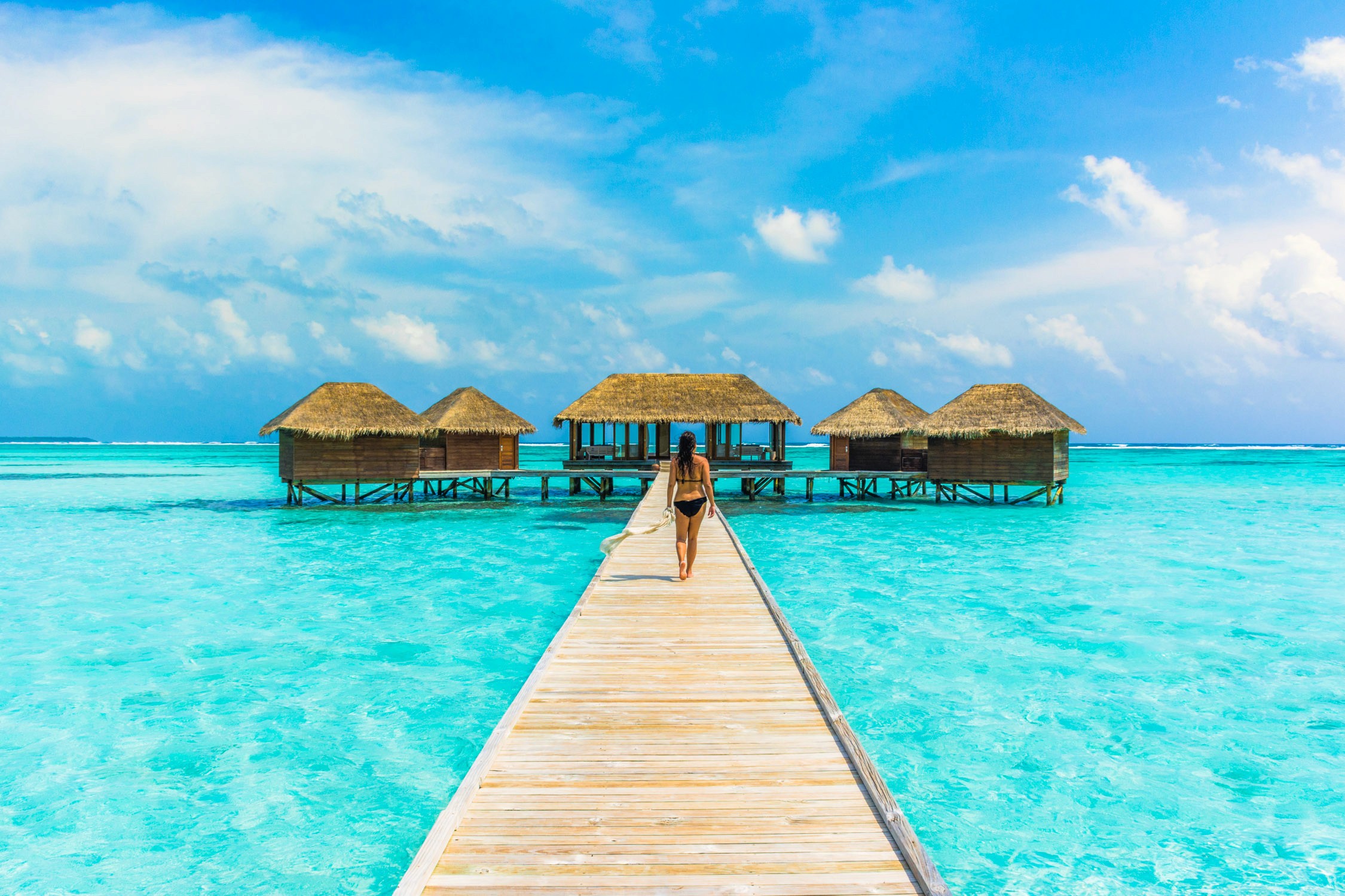 A person walks along a wooden pier toward thatched-roof overwater bungalows in turquoise tropical water under a blue sky.