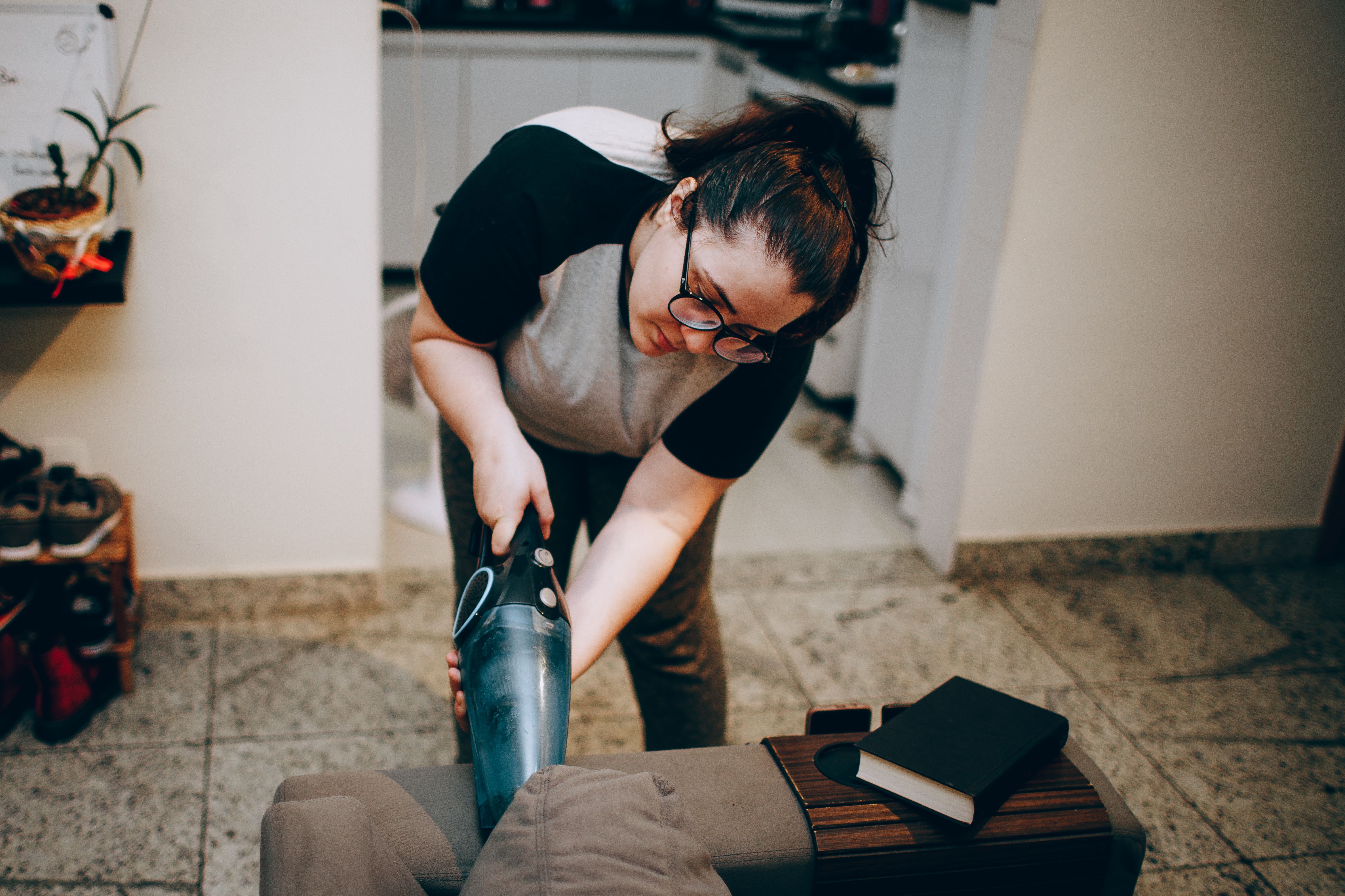 A young woman in glasses vacuums a couch cushion with a handheld vacuum, focused on one small cleaning task at a time with ADHD.
