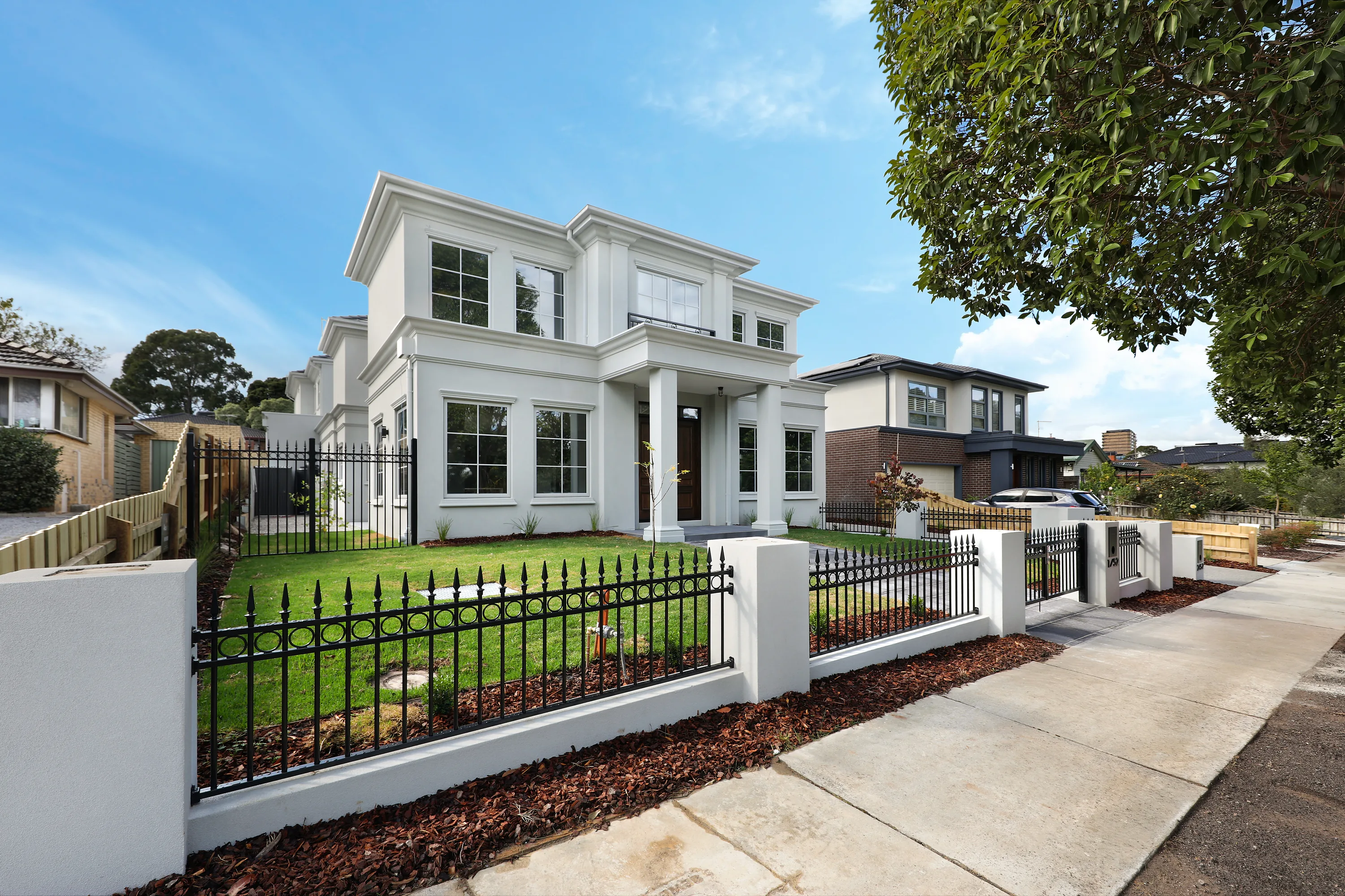 Front facade of the Glen Waverley Residence, a white-rendered two-storey classic home with moulded window surrounds, a columned portico and timber entry door behind a wrought-iron and rendered fence
