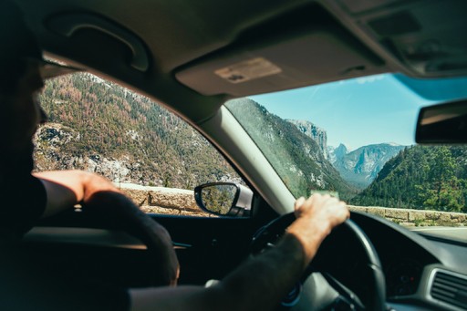 A close-up of a driver's hands on the steering wheel, with scenic mountains visible through the windshield.