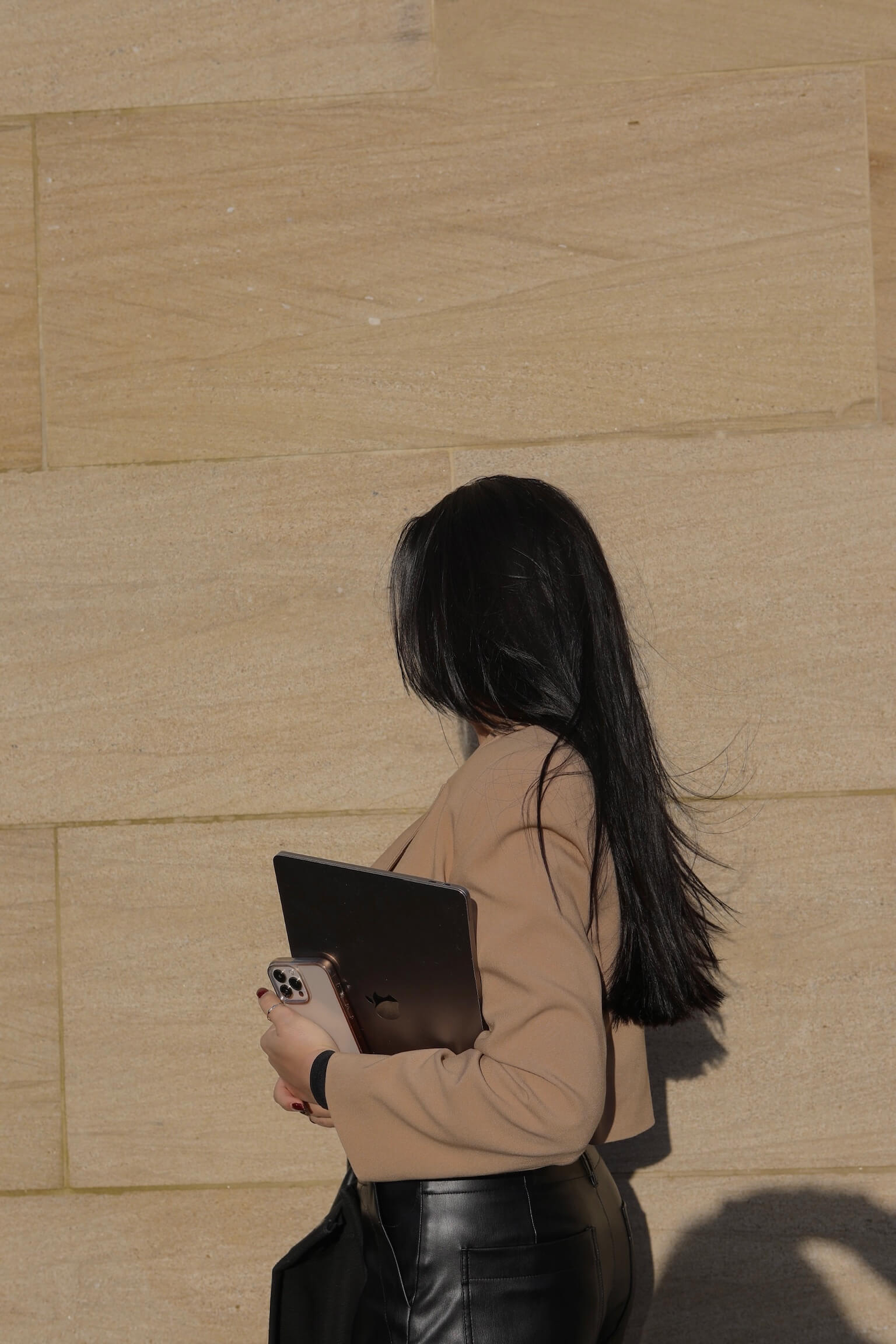 a woman with long straight dark hair, wearing a tan blazer and black leather pants, holds a laptop in her arm as she walks past a tan stone tiled wall