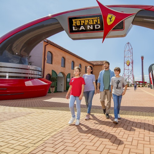 A smiling family of four walks under a large Ferrari Land entrance arch in an amusement park.
